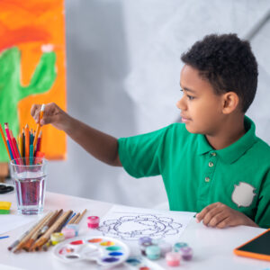 Preparation, drawing. Profile of dark-skinned involved boy in green tshirt taking out pencil from glass sitting at table preparing to draw in daylight